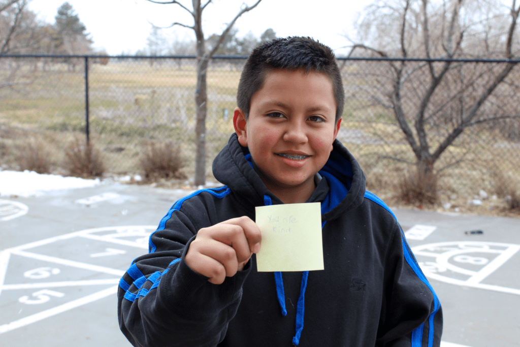 A smiling student posing outside on the Guadalupe School playground holding up a sticky note with an encouraging message written on it saying: You are kind.