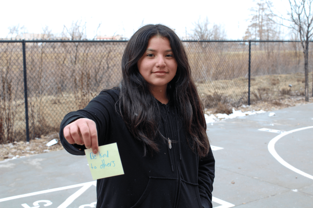 A smiling student posing outside on the Guadalupe School playground holding up a sticky note with an encouraging message written on it saying: Be kind to others.