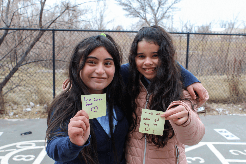 Two smiling friends posing outside on the Guadalupe School playground holding up sticky notes with encouraging messages written on them. One note says: Be a Care Bear with a heart drawn on it. The other note says: It does not matter what they say about you.