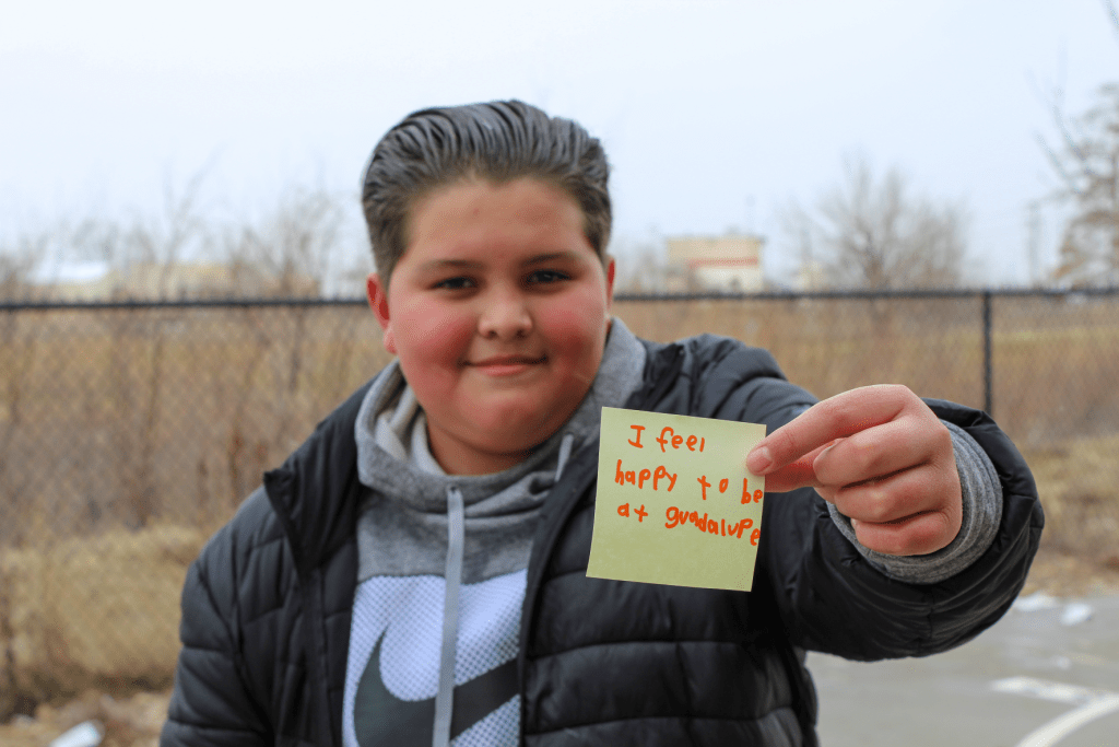 A smiling student posing outside on the Guadalupe School playground holding up a sticky note with an encouraging message written on it saying: I feel happy to be at Guadalupe.
