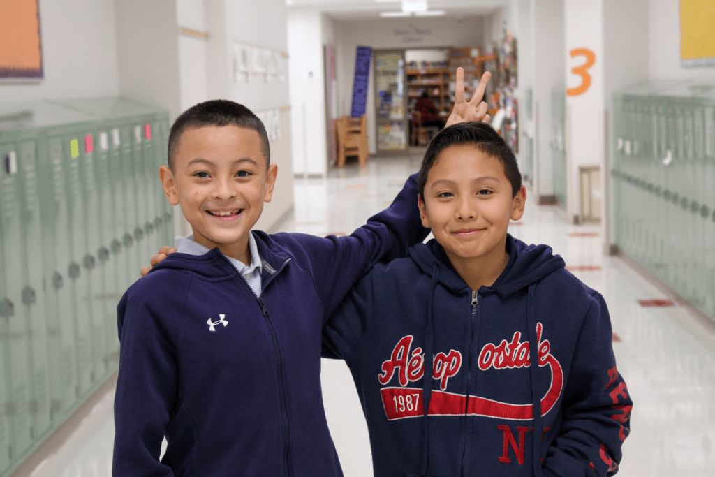 Two smiling male friends posing for a picture in the Guadalupe School hallway, one of them is playfully holding up bunny ears behind the other student's head.