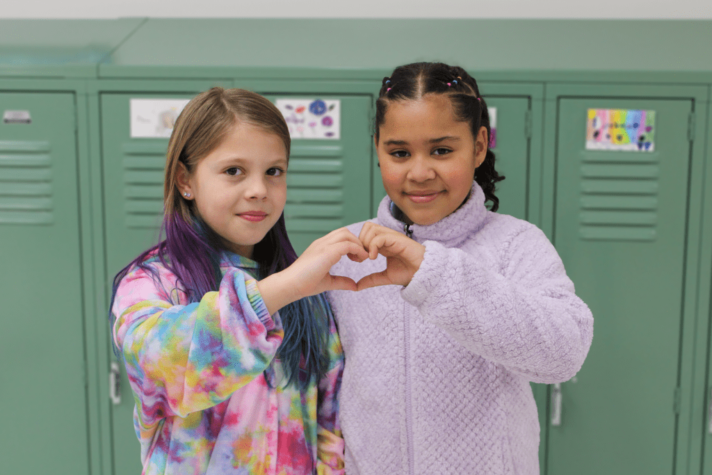 Two smiling female friends posing for a picture in front of Guadalupe School lockers, they are using their hands to make a heart together.