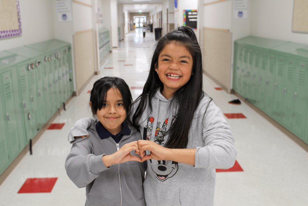 Two smiling female friends posing for a picture in the Guadalupe School hallway, they are using their hands to make a heart together.
