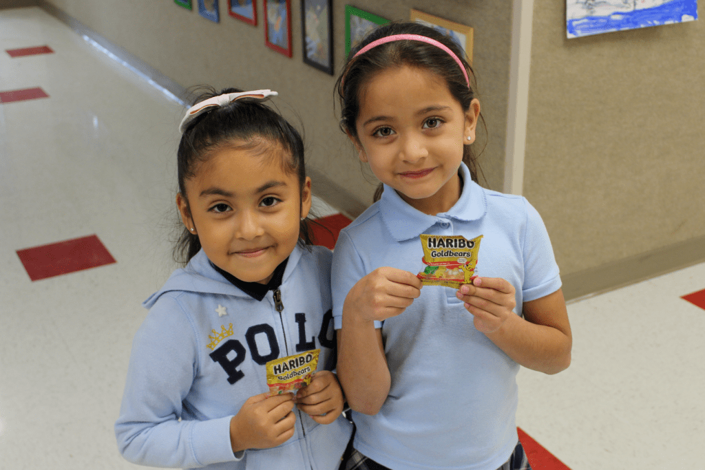 Two smiling friends posing for a picture in the Guadalupe School hallway, they are each holding their own bag of gummy bears to share during Hope Week.