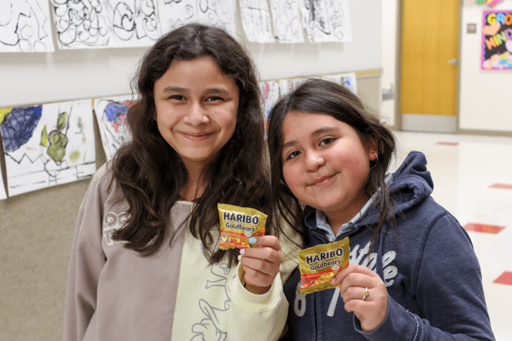 Two smiling friends posing for a picture in the Guadalupe School hallway, they are each holding their own bag of gummy bears to share during Hope Week.